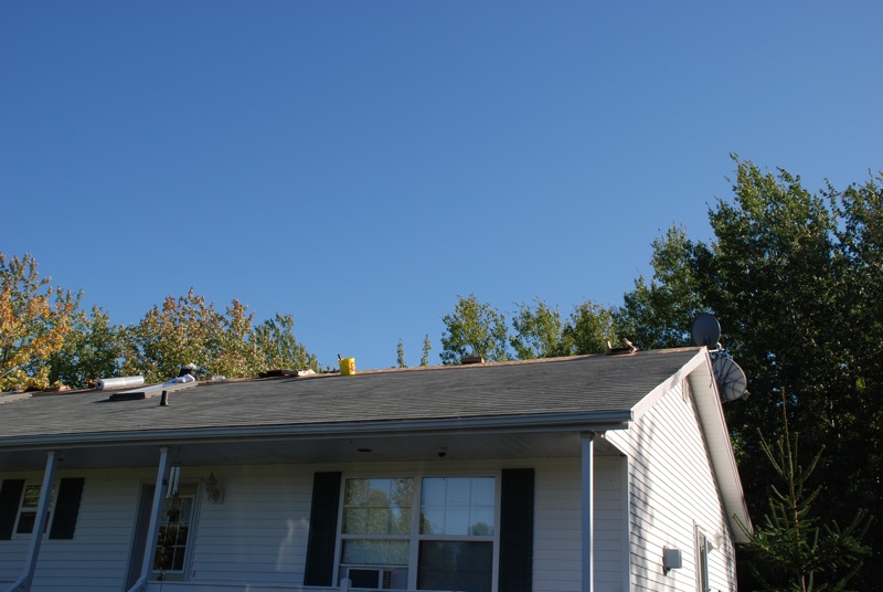 House roof with shingles, a satellite dish, and trees against a blue sky