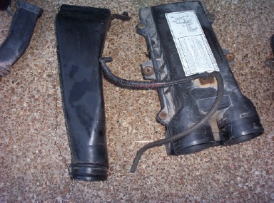 1988 Ford Bronco - Black plastic air intake components on tan carpet, showing a flat molded snorkel duct beside a larger housing with a white service/emission…