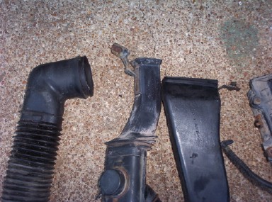 1988 Ford Bronco - Group of removed black plastic air intake ducts laid out on tan carpet, including a ribbed flexible elbow hose, a molded intake tube with a…