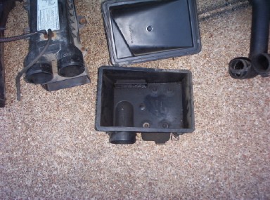 1988 Ford Bronco - Black plastic air cleaner box opened up on tan carpet, with the lid resting upside down above the empty base showing the inlet snorkel port…
