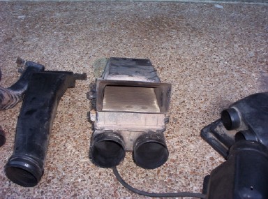 1988 Ford Bronco - Grimy plastic intake/heater box component with a rectangular open duct opening and two round outlet tubes, resting on tan carpet. Curved bl…