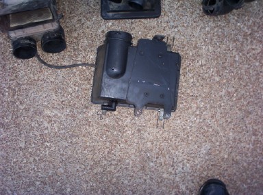 1988 Ford Bronco - Used black plastic air cleaner housing with an integrated cylindrical canister and mounting tabs, laid on tan carpet alongside other remove…