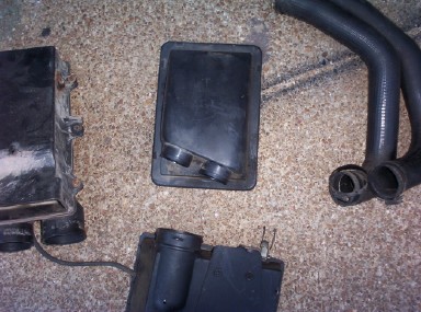 1988 Ford Bronco - Overhead shot of a disassembled air intake system on tan carpet: the air cleaner box lid lying face-up in the center, the box body at left,…