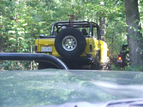 1996 Ford Bronco - Two off-road vehicles navigate a wooded trail, one yellow and one red, with a spare tire prominently displayed on the rear of the yellow ve…
