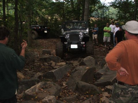 1996 Ford Bronco - A black Jeep Wrangler tackles a challenging rocky off-road course, observed by a group of onlookers.