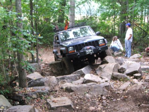 1996 Ford Bronco - A Jeep Cherokee XJ tackles a challenging off-road course with large rocks and trees surrounding the path.