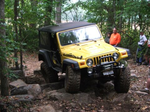 1996 Ford Bronco - A yellow Jeep Wrangler Rubicon tackles a challenging off-road course with large rocks and trees surrounding it.