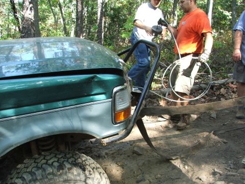 1996 Ford Bronco - Men are using a winch to pull a damaged green pickup truck out of a wooded area.