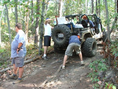 1996 Ford Bronco - People work on an off-road Jeep with large tires on a wooded trail.
