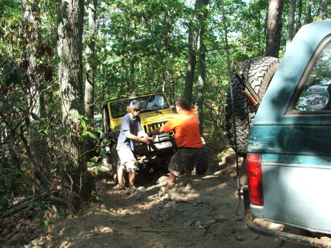 1996 Ford Bronco - Two people attempt to free a yellow Jeep from a muddy trail while a green SUV waits behind.
