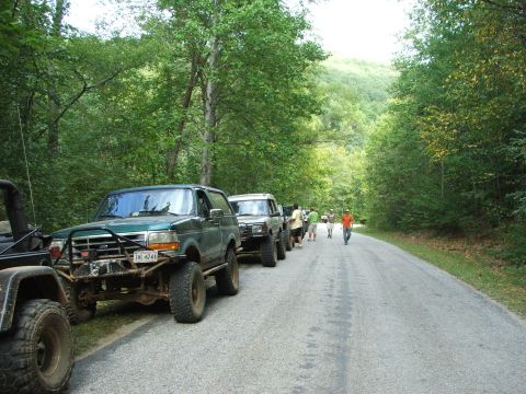 1996 Ford Bronco - A line of off-road vehicles, including a 1996 Ford Bronco, are parked on the side of a tree-lined road.