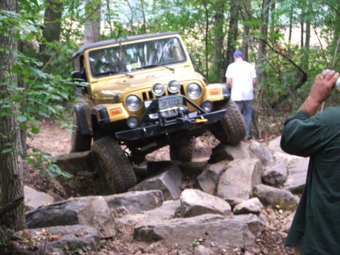 1996 Ford Bronco - A yellow Jeep Wrangler is shown off-roading over a challenging rock obstacle in a wooded area.