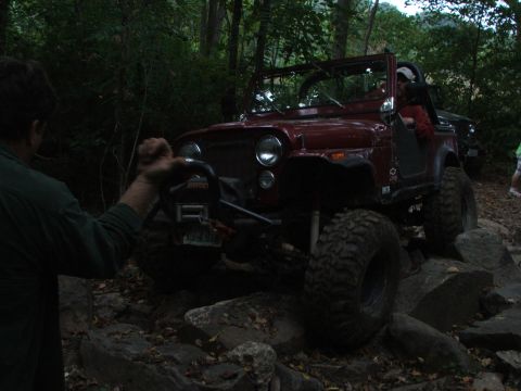 1996 Ford Bronco - A red off-road jeep is shown navigating a challenging rocky trail, with a person's hand visible on the winch.