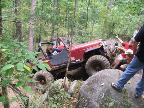 1996 Ford Bronco - A red Jeep Wrangler is shown off-roading on a rocky trail with several people observing and assisting.