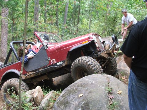 1996 Ford Bronco - A red Jeep Wrangler is shown off-roading through a challenging rocky course with spectators observing the action.