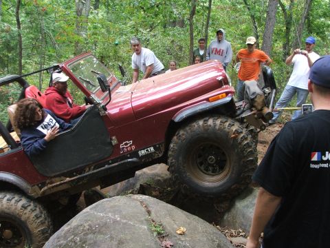 1996 Ford Bronco - A red Jeep Wrangler tackles a challenging off-road course with spectators watching.
