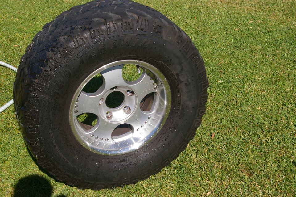 Close-up of a large off-road tire on a silver rim, sitting on green grass. Some water droplets are visible on the tire