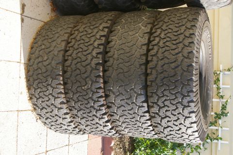 1986 Ford Bronco - Four off-road tires are stacked on their sides on a paved surface.