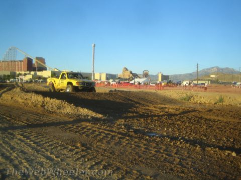 1990 Ford Bronco - yellow off-road pickup truck drives along a dirt berm with mountains, power lines and a fairground in the background.
