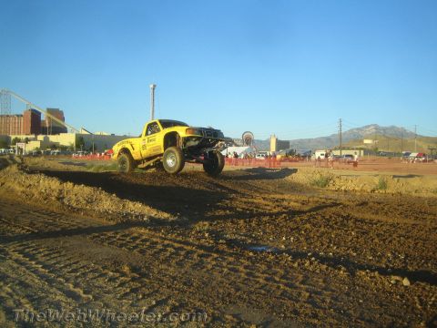 1990 Ford Bronco - yellow off-road pickup truck launches off a dirt berm with mountains and a fairground in the background.
