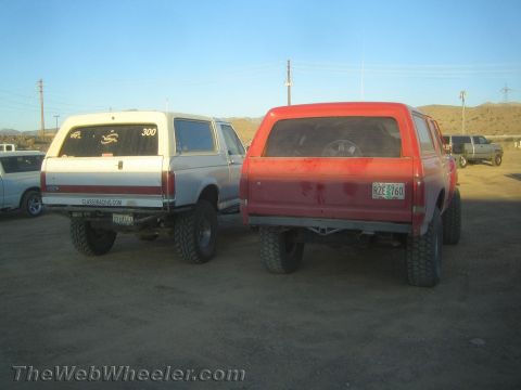 1990 Ford Bronco - white and a red lifted full-size SUV parked rear-to-camera on a dusty desert lot with power lines behind.