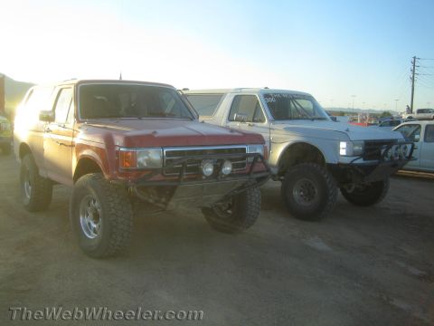 1990 Ford Bronco - red lifted full-size SUV with a tubular brush guard and auxiliary lights parked next to a white SUV on a dirt lot.