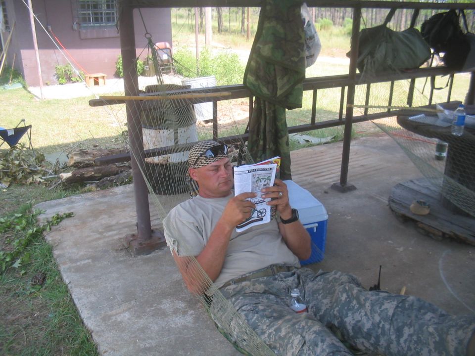 Man in Army camo reclining in a hammock reading at an outdoor camp