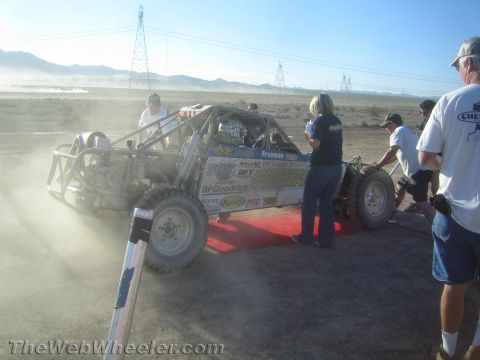1990 Ford Bronco - tube-chassis off-road race buggy sits on a red ramp surrounded by crew members in a dusty desert staging area.