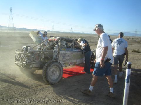 1990 Ford Bronco - tube-frame off-road race buggy on a red ramp as crew members walk past amid desert dust and power lines.