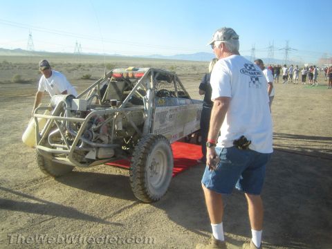 1990 Ford Bronco - Crew members tend to a tube-frame off-road race buggy on a red ramp in a dusty desert pit area.