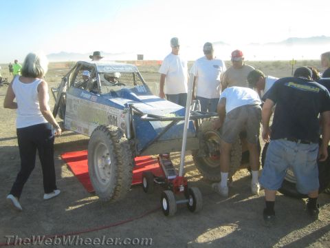 1990 Ford Bronco - Crew members raise a blue tube-frame off-road buggy on a floor jack to change a rear tire in the desert.