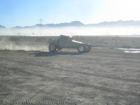 1990 Ford Bronco - off-road race buggy speeds across a flat desert course raising dust beneath transmission towers and mountains.