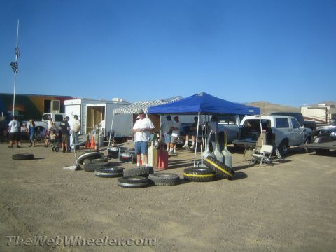 1990 Ford Bronco - desert race pit area with a blue canopy, RVs, trucks and rows of stacked tires under a clear sky.