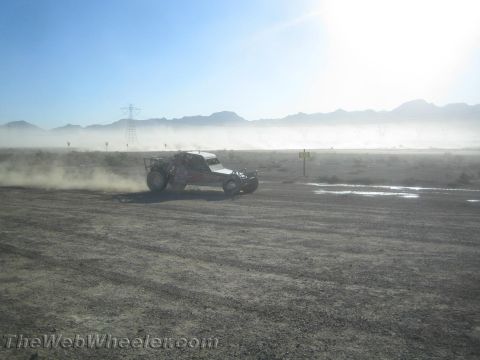 1990 Ford Bronco - off-road race buggy speeds across a dusty desert course backlit by bright sun with mountains in the distance.