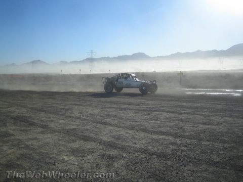 1990 Ford Bronco - white off-road race buggy kicks up dust as it drives across a flat desert course under hazy backlight.