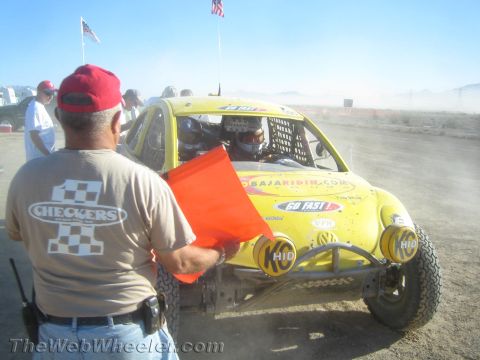 1990 Ford Bronco - man in a Checkers club shirt holds an orange flag beside a yellow off-road race buggy in the dusty desert.