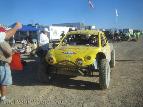 1990 Ford Bronco - yellow off-road race buggy numbered 500 sits in a desert pit lane lined with trailers, canopies and crew.