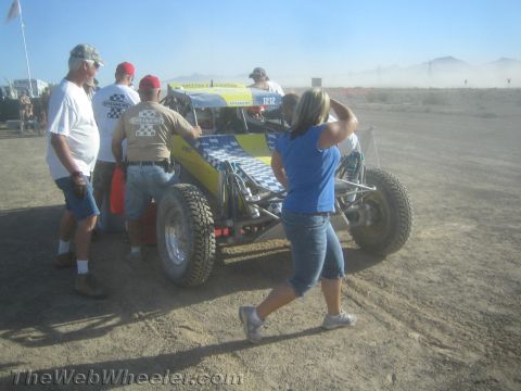 1990 Ford Bronco - Crew members gather around a blue and yellow checkered tube-frame off-road buggy in a dusty desert pit.