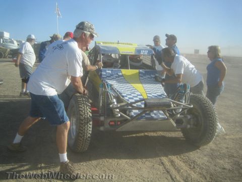 1990 Ford Bronco - Several crew members push a blue and yellow checkered tube-frame off-road buggy across a dusty desert pit.