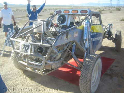 1990 Ford Bronco - number 212 tube-frame off-road race buggy sits on a red ramp as a man cheers with raised arms in the desert.