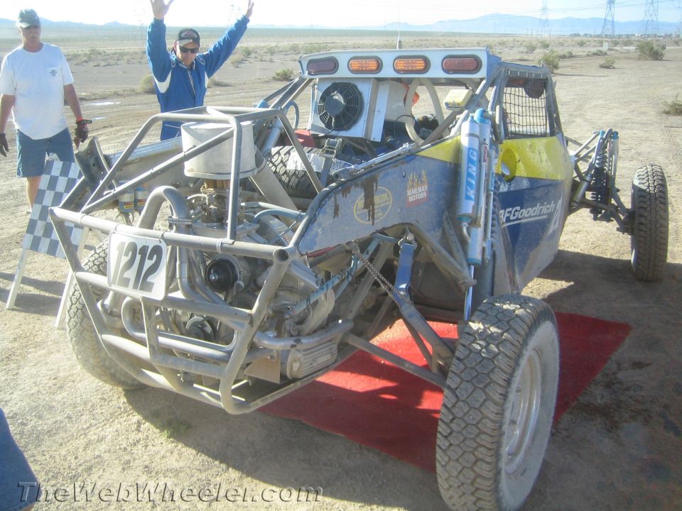 Number 212 off-road race buggy on a red ramp with celebrating crew in desert