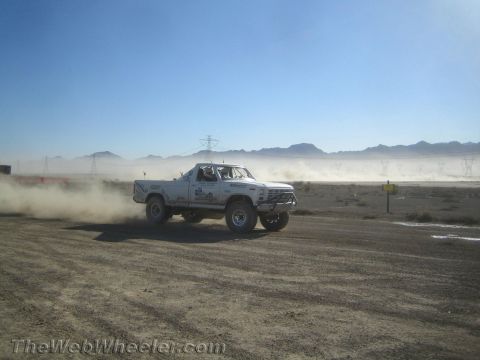 1990 Ford Bronco - white off-road race pickup speeds across a dusty desert course with power-line towers and mountains in the background.