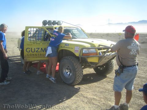 1990 Ford Bronco - Crew and spectators gather around a yellow off-road race truck marked Guzman Construction at a dusty desert pit area.