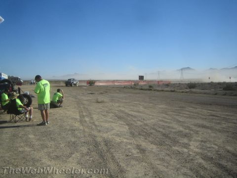 1990 Ford Bronco - People in bright green shirts sit and stand watching a distant vehicle raise dust along an orange-fenced desert race course.