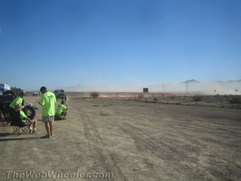 1990 Ford Bronco - People in green shirts watch a dark pickup kick up dust along an orange-fenced desert off-road course.