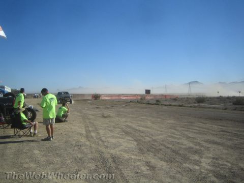1990 Ford Bronco - dark off-road pickup raises dust on a desert course as spectators in green shirts look on near power-line towers.