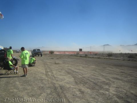 1990 Ford Bronco - blue-and-white off-road pickup drives along an orange-fenced desert course as spectators watch from the dirt staging area.
