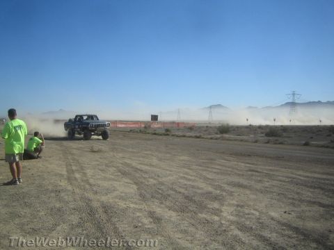 1990 Ford Bronco - dark off-road race pickup throws up dust crossing a desert course as a spectator watches under a clear blue sky.
