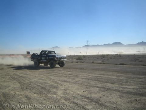 1990 Ford Bronco - Race truck number 300 speeds across a dusty desert course with mountains and power lines behind it.