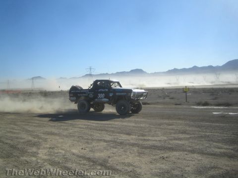 1990 Ford Bronco - Blue race truck number 300 drives along a dusty desert course beneath power-line towers and distant mountains.